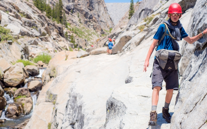 A student wearing a helmet braces themself on a rock wall while making their way across narrow rocky terrain. There is a stream below them.